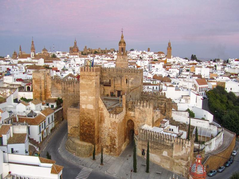 A medieval fortress gate glowing over the white rooftops of Carmona at dusk, Seville — explore the Campiña de Carmona with Andaluciatours.