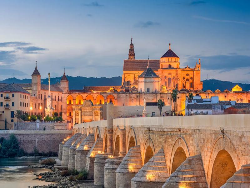 Córdoba's Roman Bridge and the floodlit Mosque-Cathedral at dusk over the Guadalquivir — discover Córdoba with Andaluciatours.