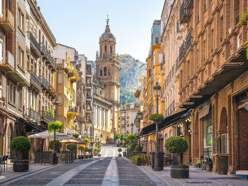 A paved old-town street lined with elegant townhouses and cafés leading to Jaén's Renaissance cathedral tower, with the Santa Catalina hill beyond — explore the Jaén metropolitan area with Andaluciatours.